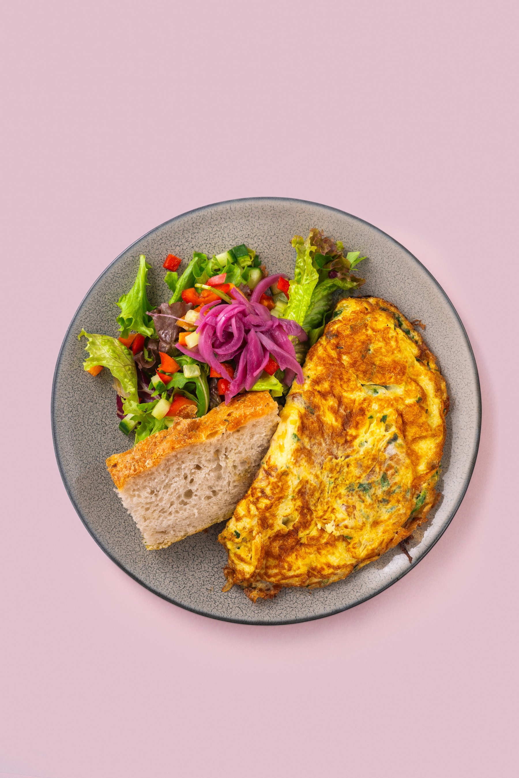 Tallerken med omelett, salat og brød mot lys rosa bakgrunn.