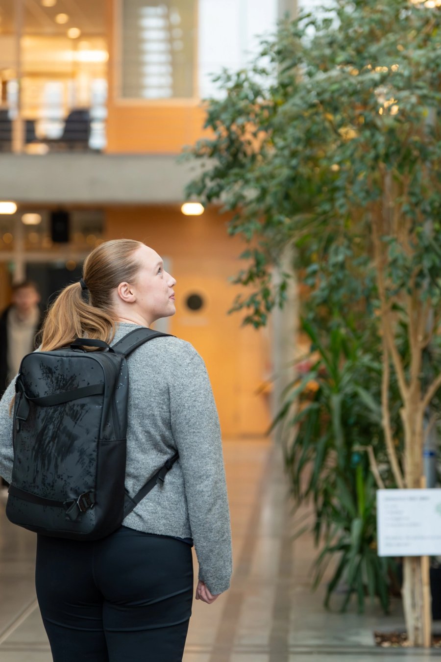 Student med ryggsekk går gjennom et lyst innendørsområde med grønne planter i et NTNU-bygg.