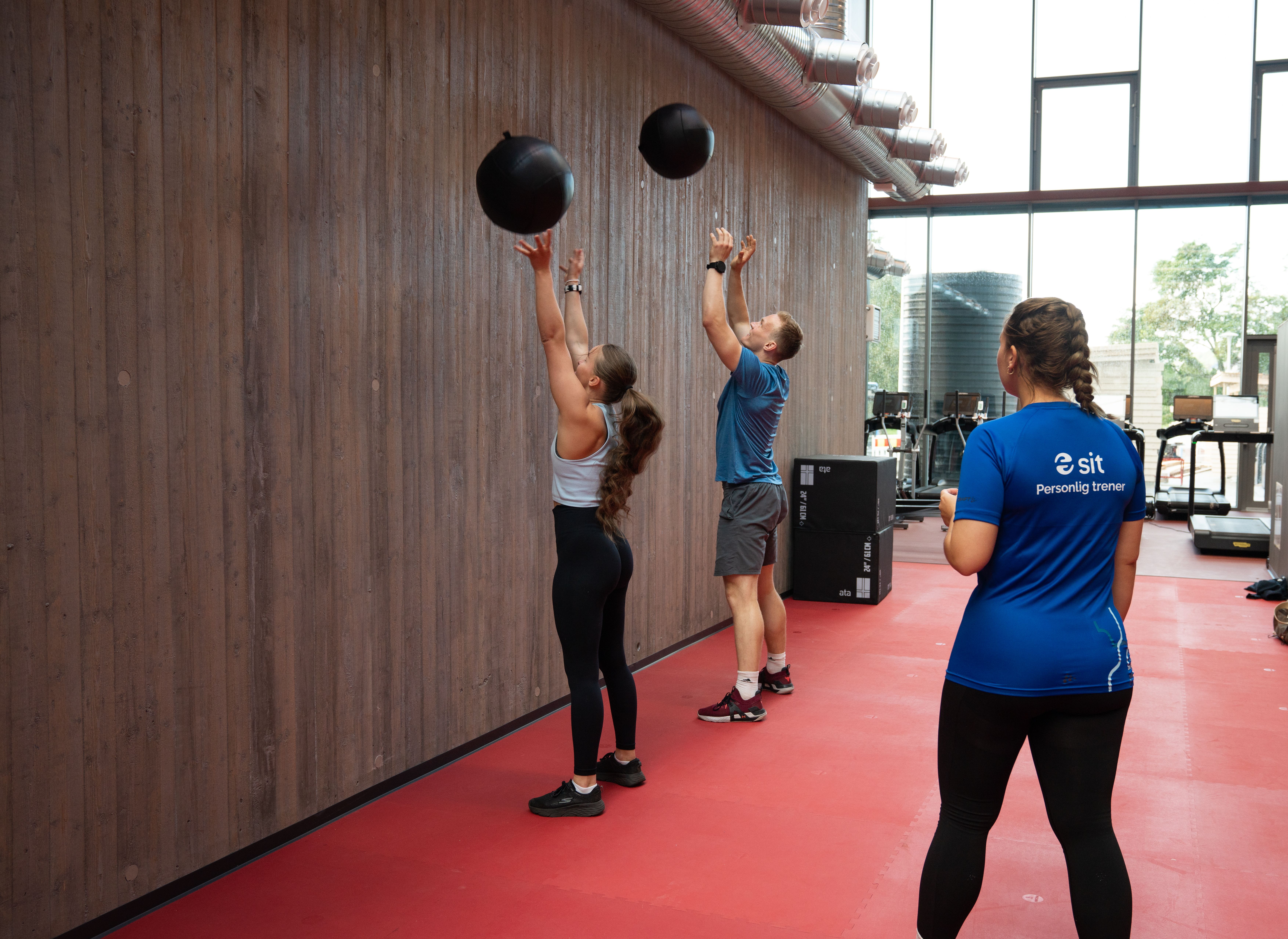 To personer som kaster medisinball høyt opp på veggen, under tilsyn av en personlig trener. 