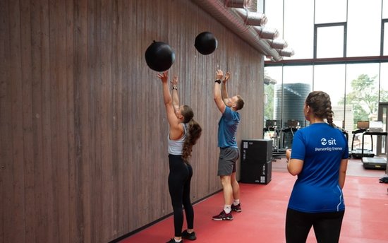 To personer som kaster medisinball høyt opp på veggen, under tilsyn av en personlig trener.