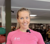 Portrett av Trine, leder for trening og tilbud i Sit Trondheim. Hun smiler mot kamera og står innendørs i et treningsmiljø.