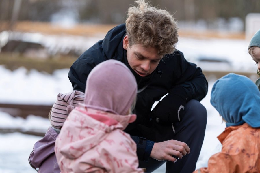 Photo of an educator in a black jacket talking to three children dressed in winter clothes out in the snow. The children are listening attentively.