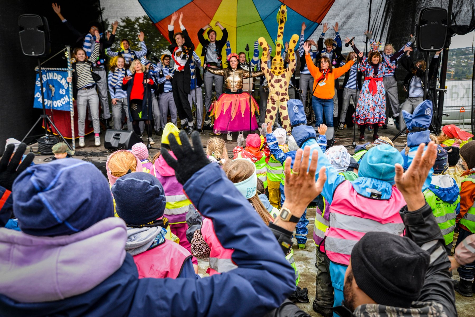 En gruppe artister i fargerike kostymer og stripete gensere opptrer på scenen foran et publikum av barn i regntøy og refleksvester, som rekker hendene i været.