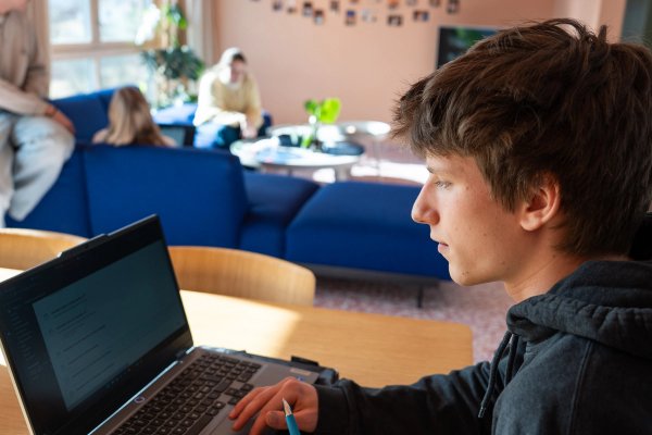 En mann sitter ved en PC og ser på skjermen. I bakgrunnen sitter to kvinner i en sofa