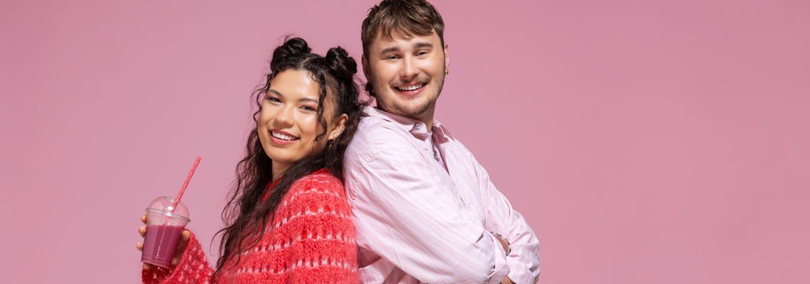 Bilde av to studenter, en mann og en kvinne som står med ryggen mot hverandre og smiler. Kvinnen holder en smoothe i hånda. 