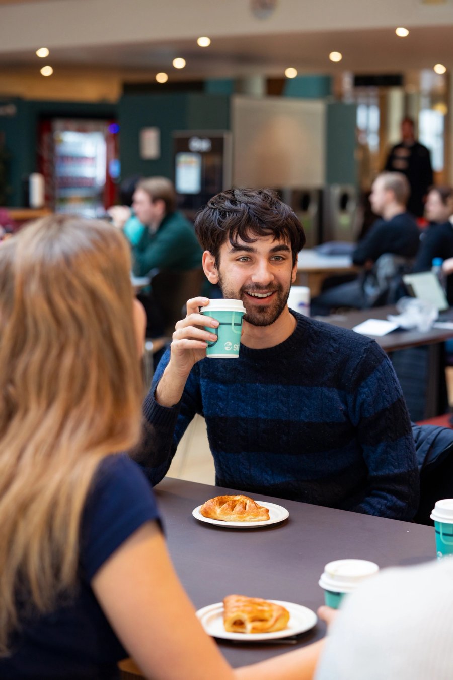 Student sitter i kantine og drikker kaffe mens han snakker med en medstudent.