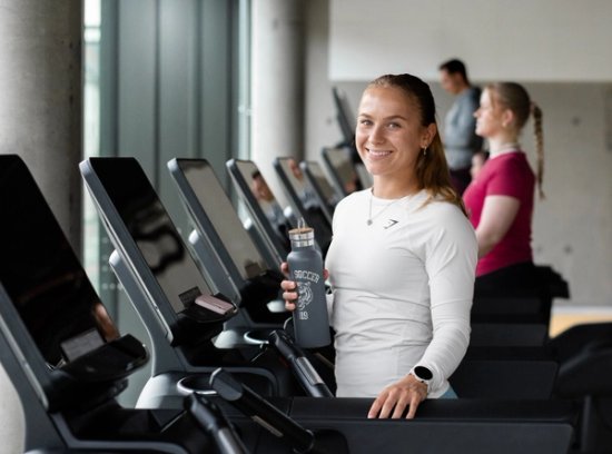 Foto av en blid jente som står på en tredemølle og holder en vannflaske. 