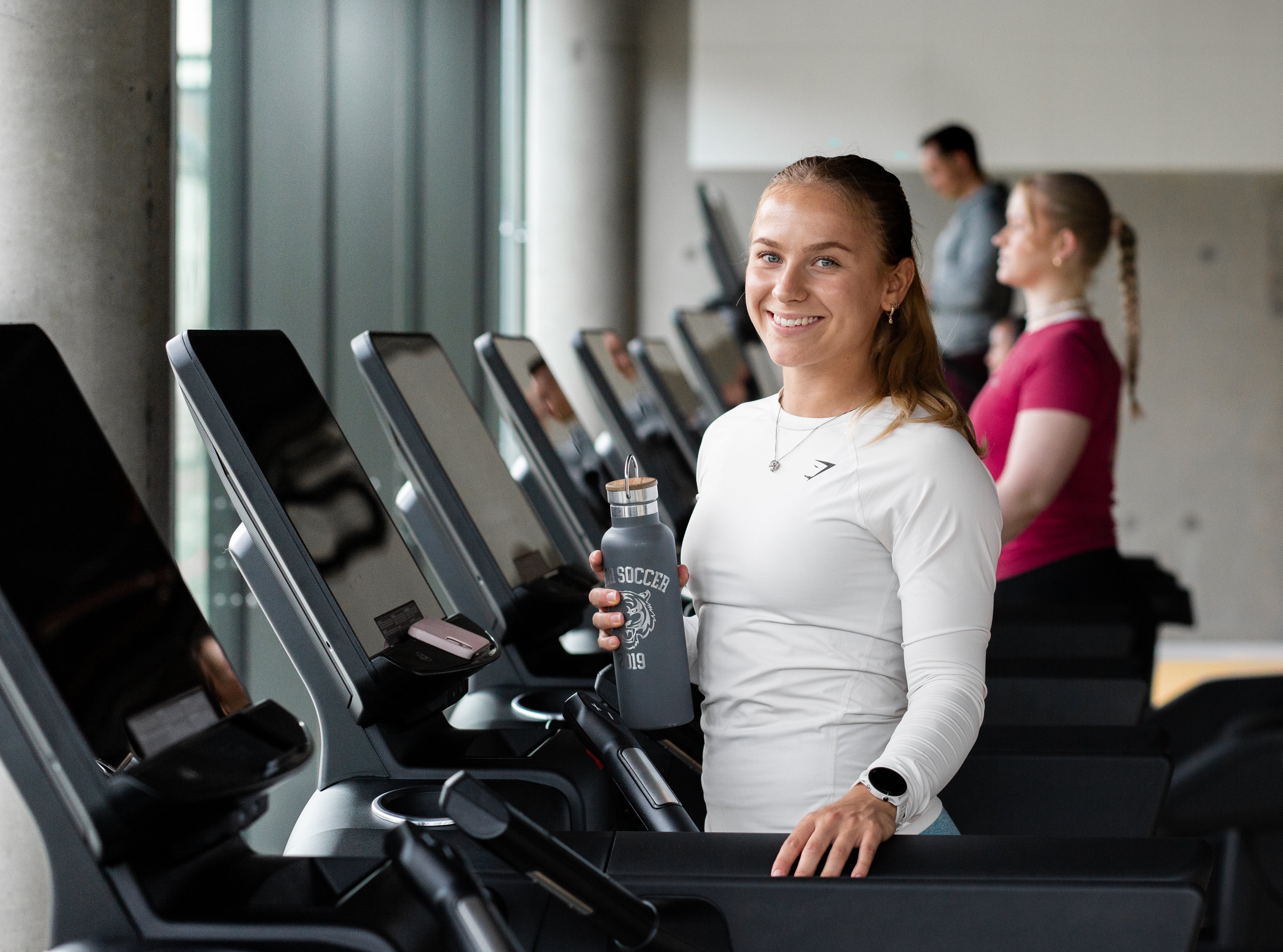 Foto av en blid jente som står på en tredemølle og holder en vannflaske.