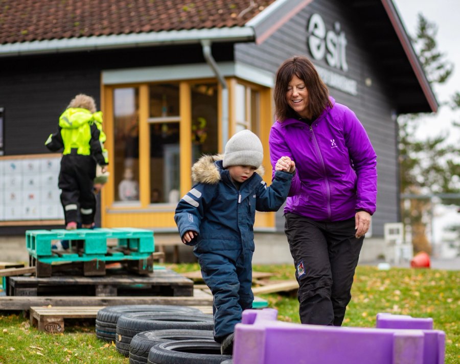 A kindergarten staff member holds a child’s hand as they balance on a car tyre outside. In the background, another child is playing on a platform made of pallets in front of a building marked with "Sit".