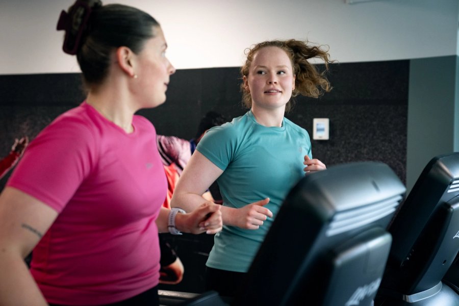 Two girls running on a treadmill next to each other and looking at each other.