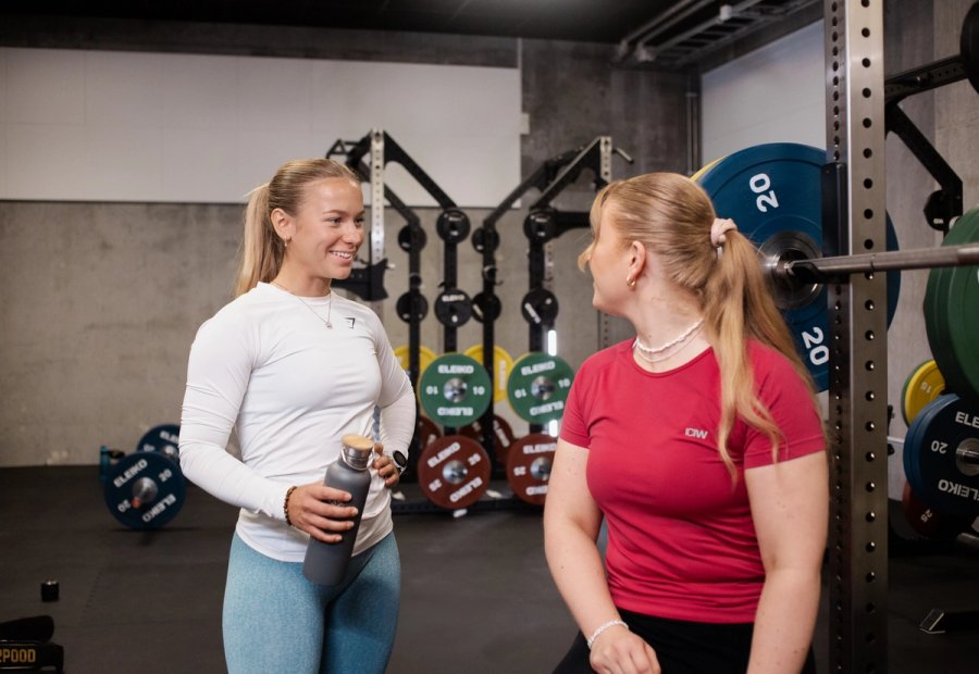 To personer ved en vektstang på et treningssenter. Den ene sitter ved stangen, den andre står med drikkeflaske. Fargede vektskiver er synlige i bakgrunnen.
