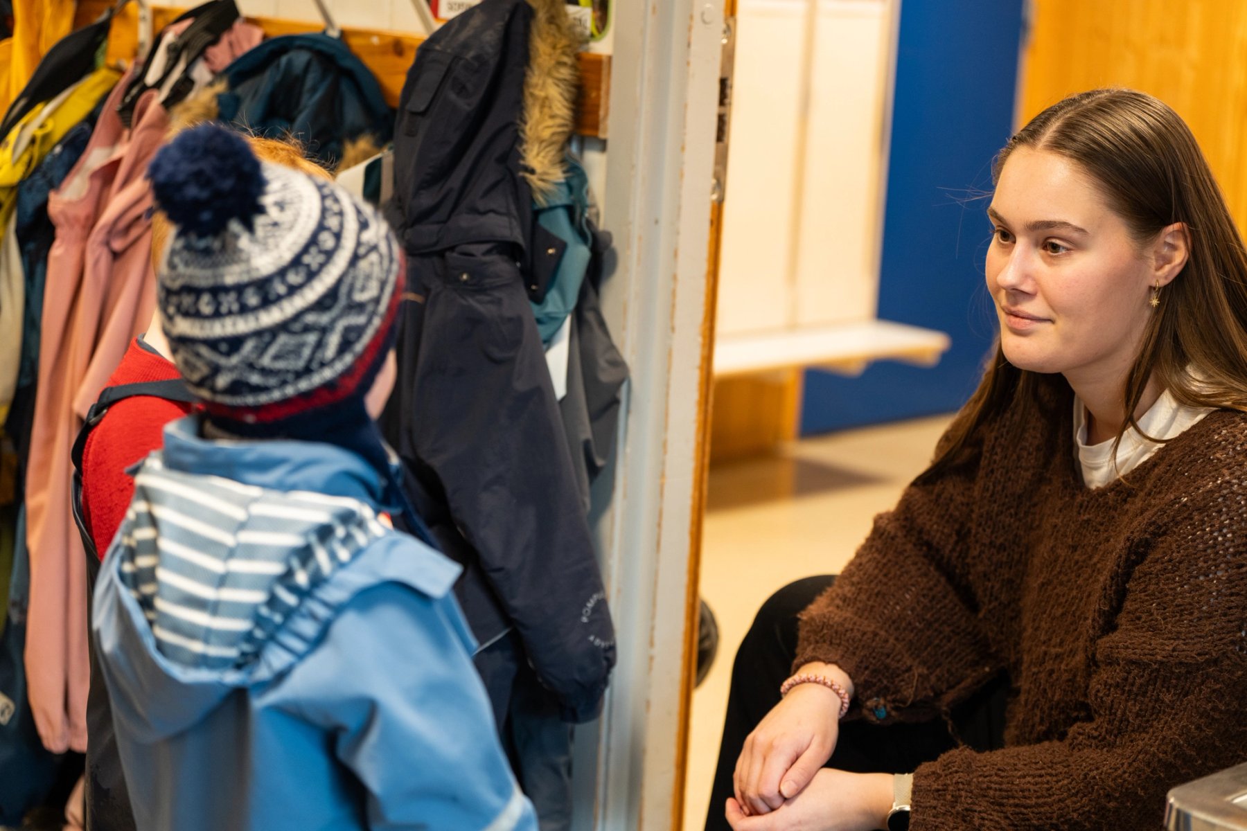 Foto av en barnehageansatt sitter på huk og snakker med et barn i garderoben. Barnet har på seg en blå jakke og en lue med mønster, mens flere jakker henger på knagger i bakgrunnen.