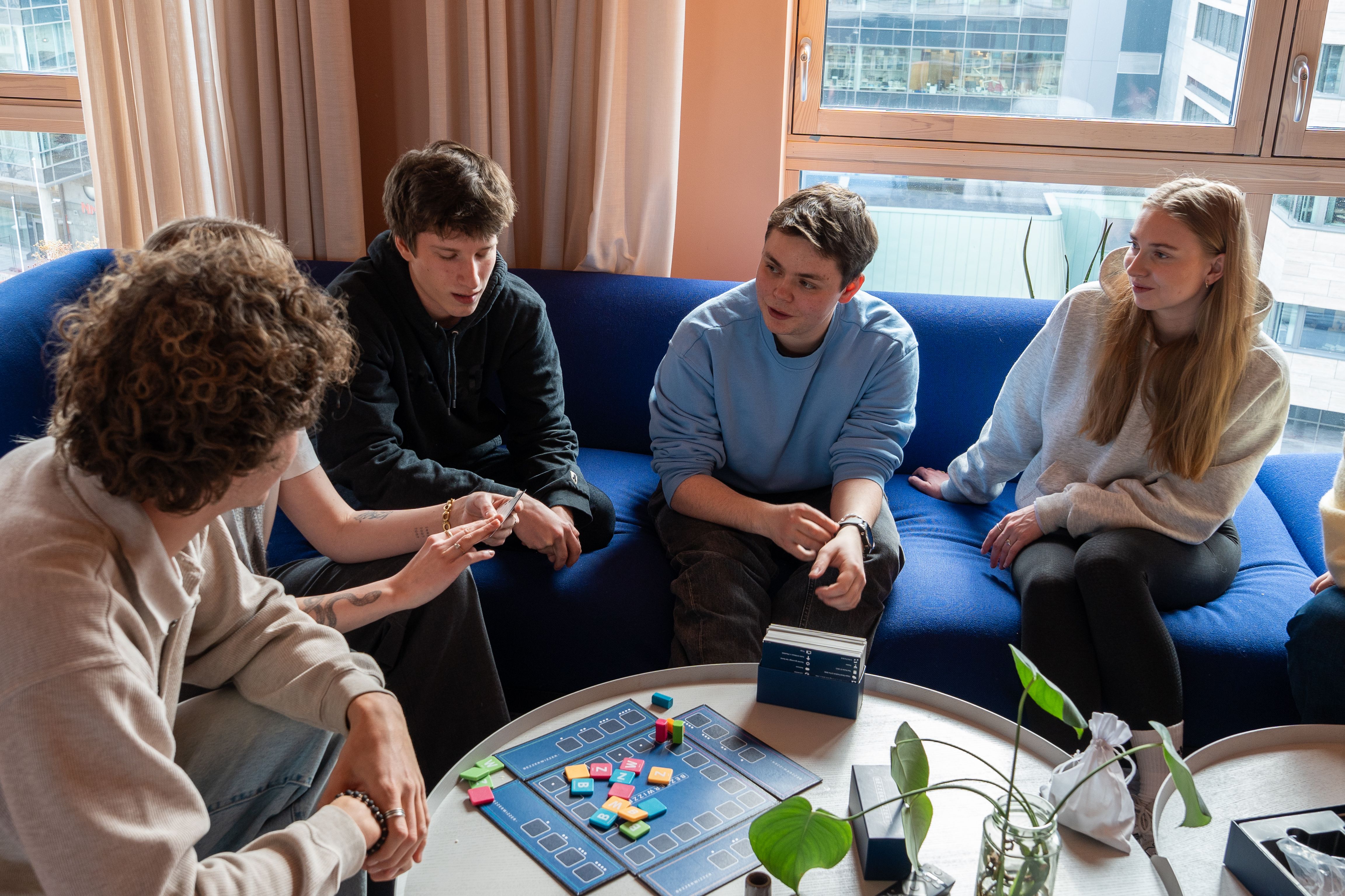 Flere studenter sitter sammen i en sofa i et felles oppholdsrom og spiller brettspill rundt et bord.