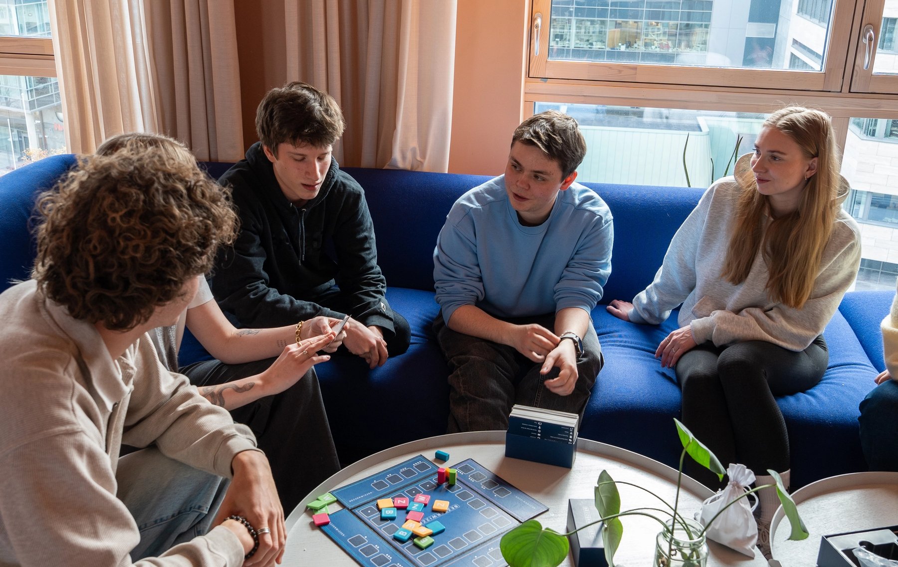 Flere studenter sitter sammen i en sofa i et felles oppholdsrom og spiller brettspill rundt et bord.
