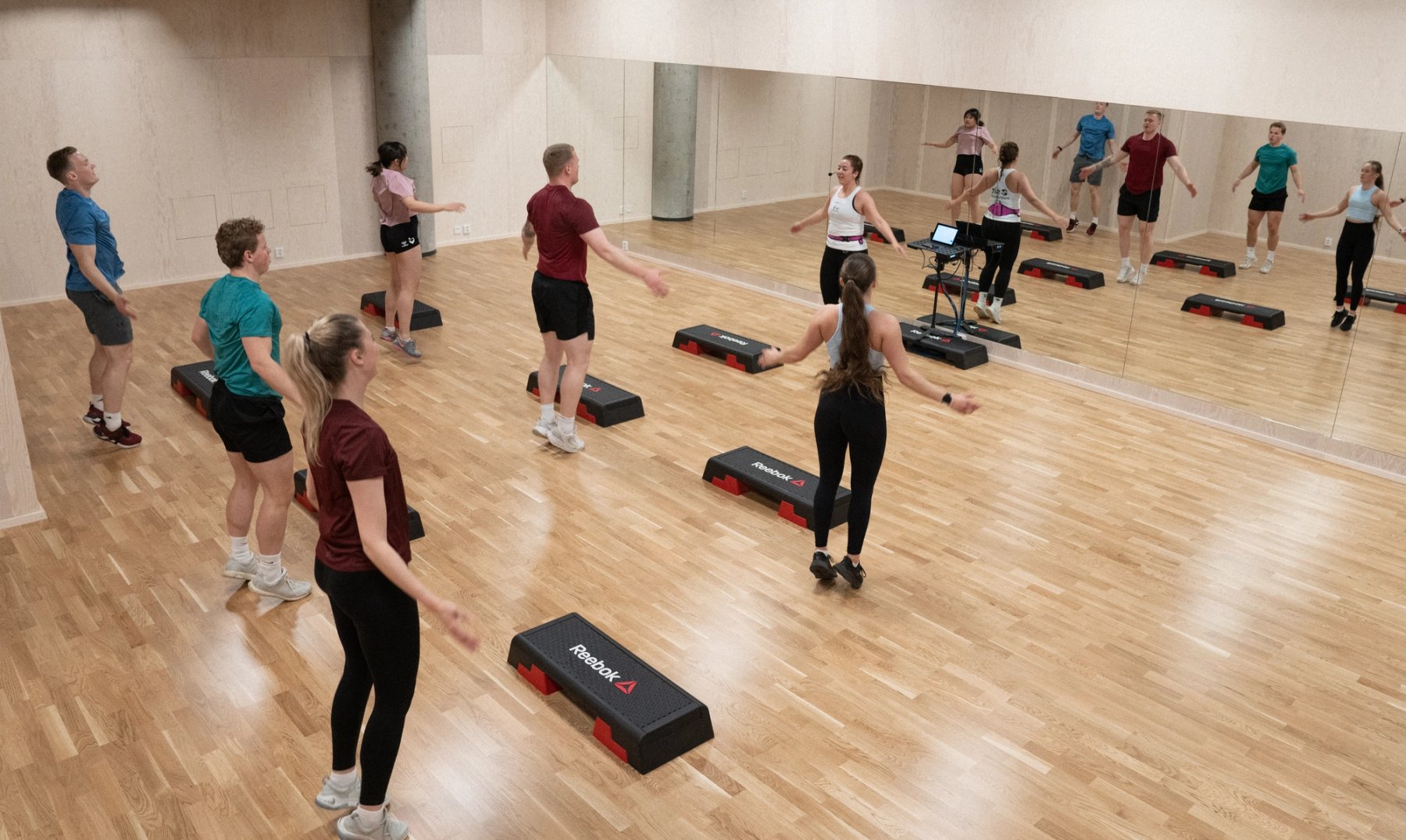Personer som trener med step i en lys gruppetreningssal med parkett på gulvet og speil langs ene veggen.