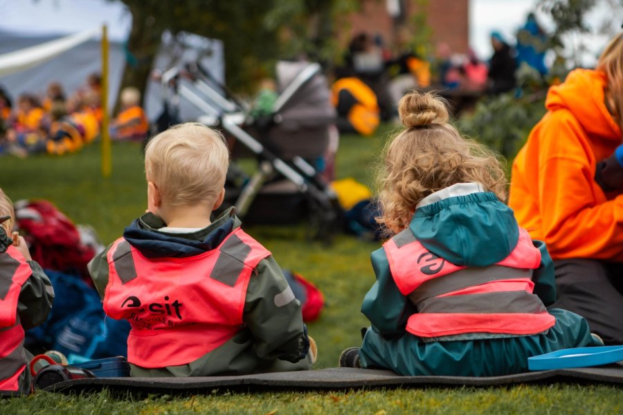 Photo of two children wearing reflective vests sitting on the grass during an outdoor activity. In the background, more children and adults are gathered, with prams parked nearby. The atmosphere is lively, filled with play and togetherness.