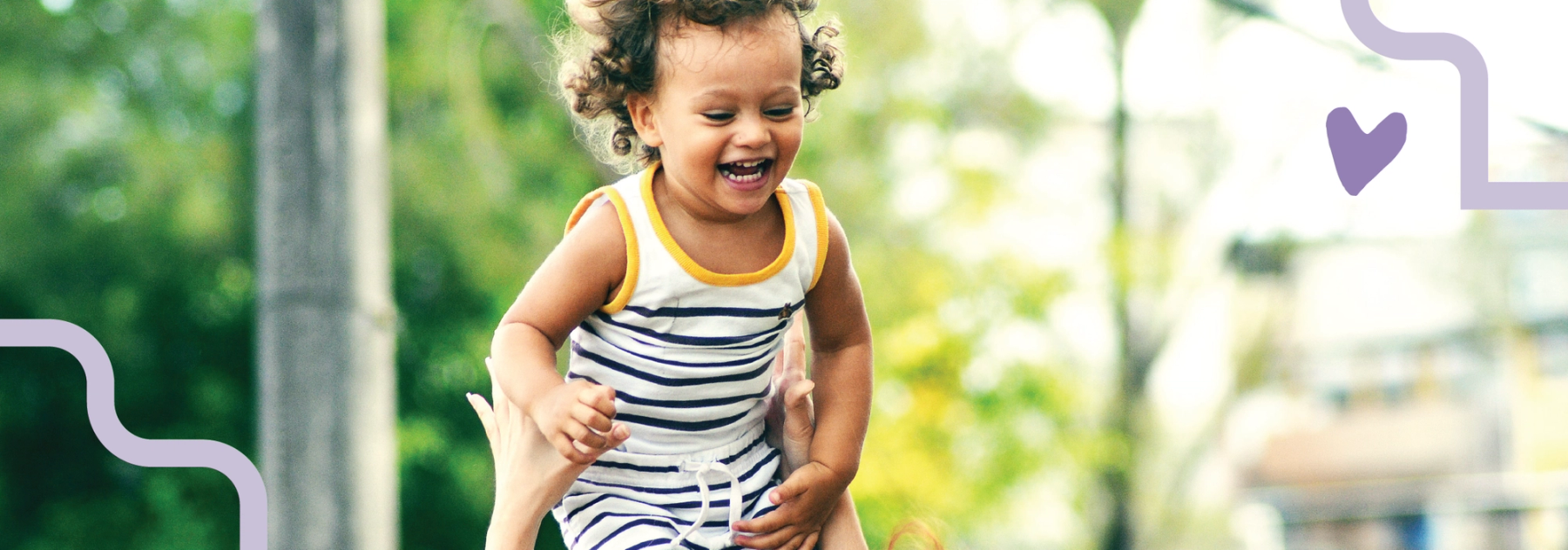 Et lite barn ler og blir løftet opp utendørs, omgitt av grønne omgivelser.