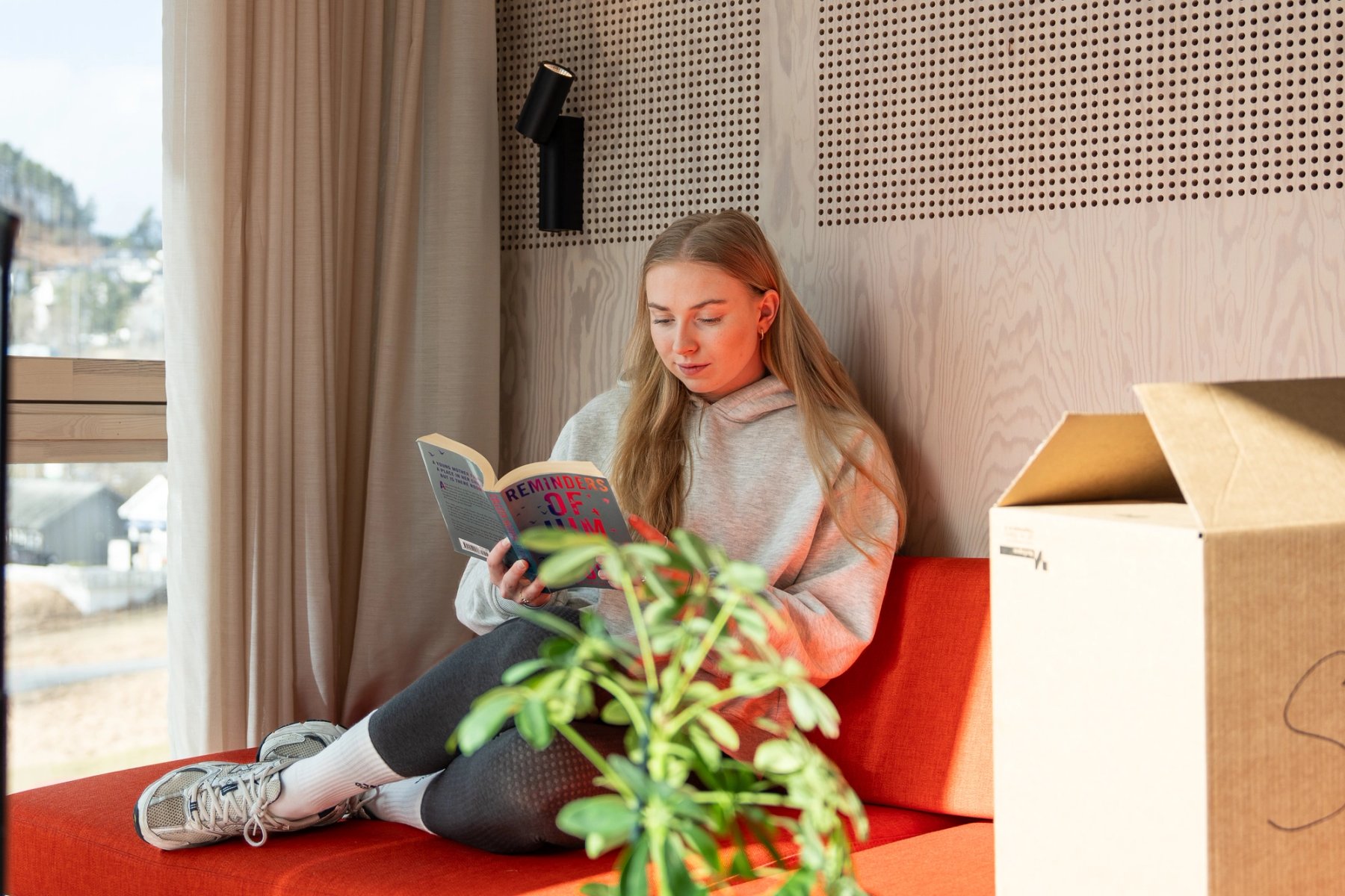 Student sitter i en møblert studentbolig og leser bok, med flyttekasse ved siden av.