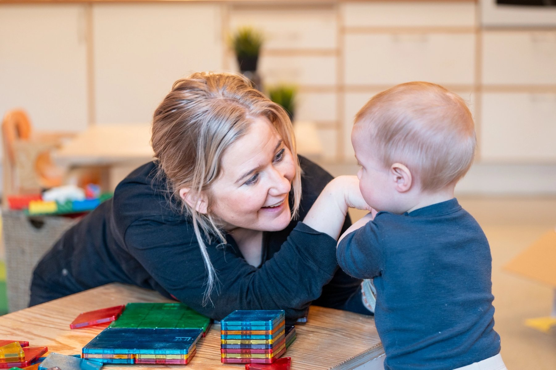 A staff member smiles warmly at a small child standing by a table. They share eye contact and a present moment together in the play environment. Colourful building bricks are on the table.