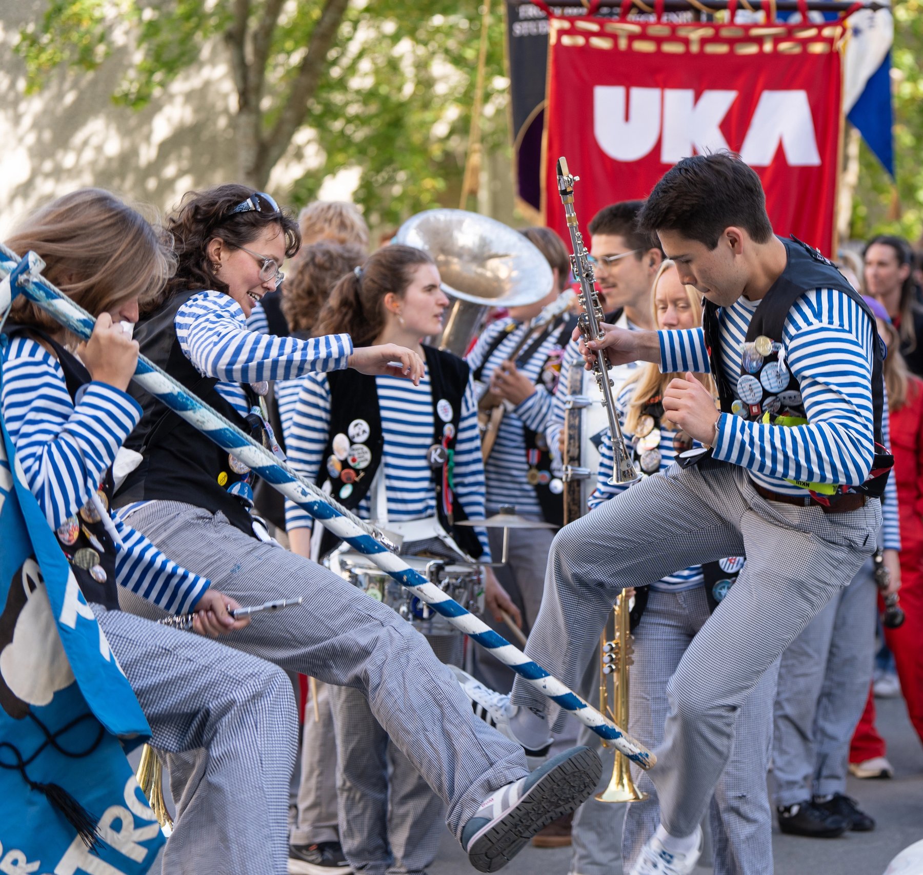 Tre musikanter i Strindens Promenade Orchester hopper og spiller musikk under immatrikulering ved NTNU i Trondheim. Det er også en rød UKA-logo i bakgrunnen