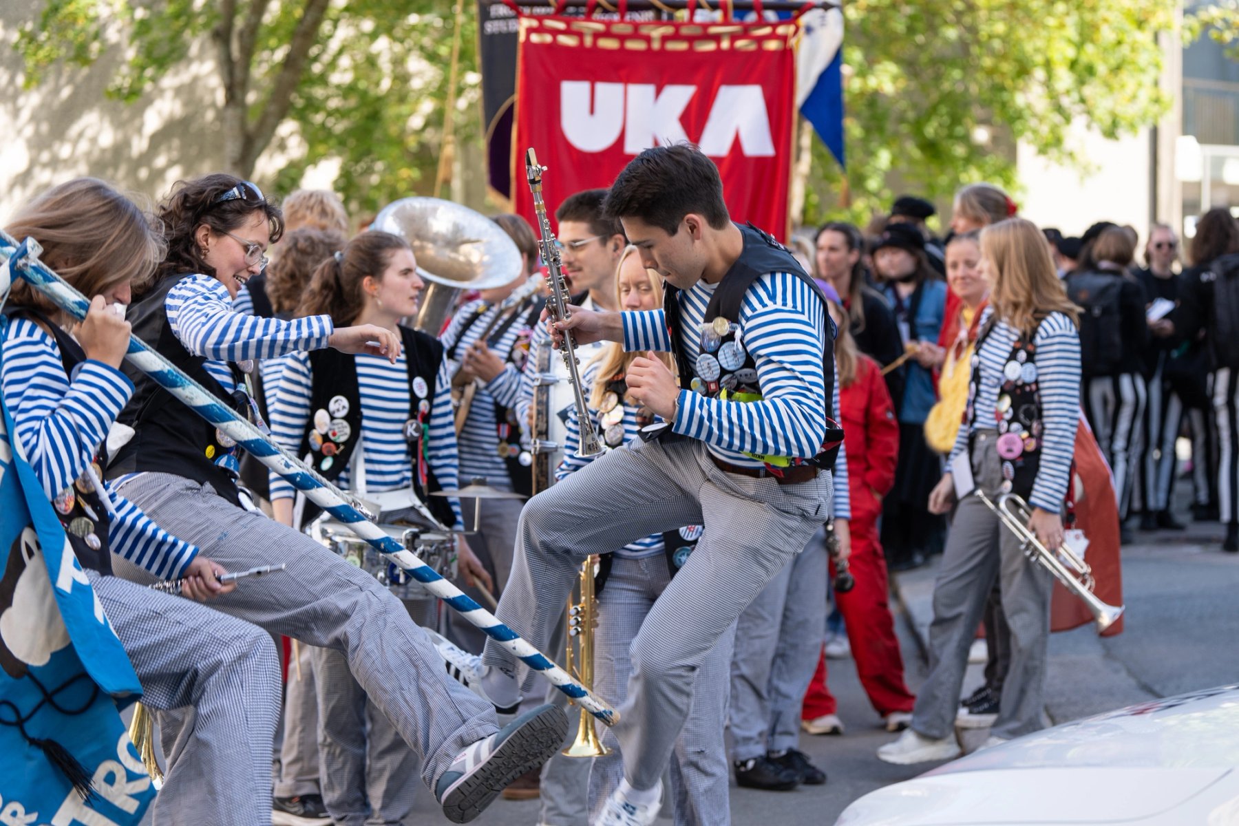 Three musicians from Strindens Promenade Orchester jump and play music during the matriculation ceremony at NTNU in Trondheim. A red UKA logo is visible in the background.