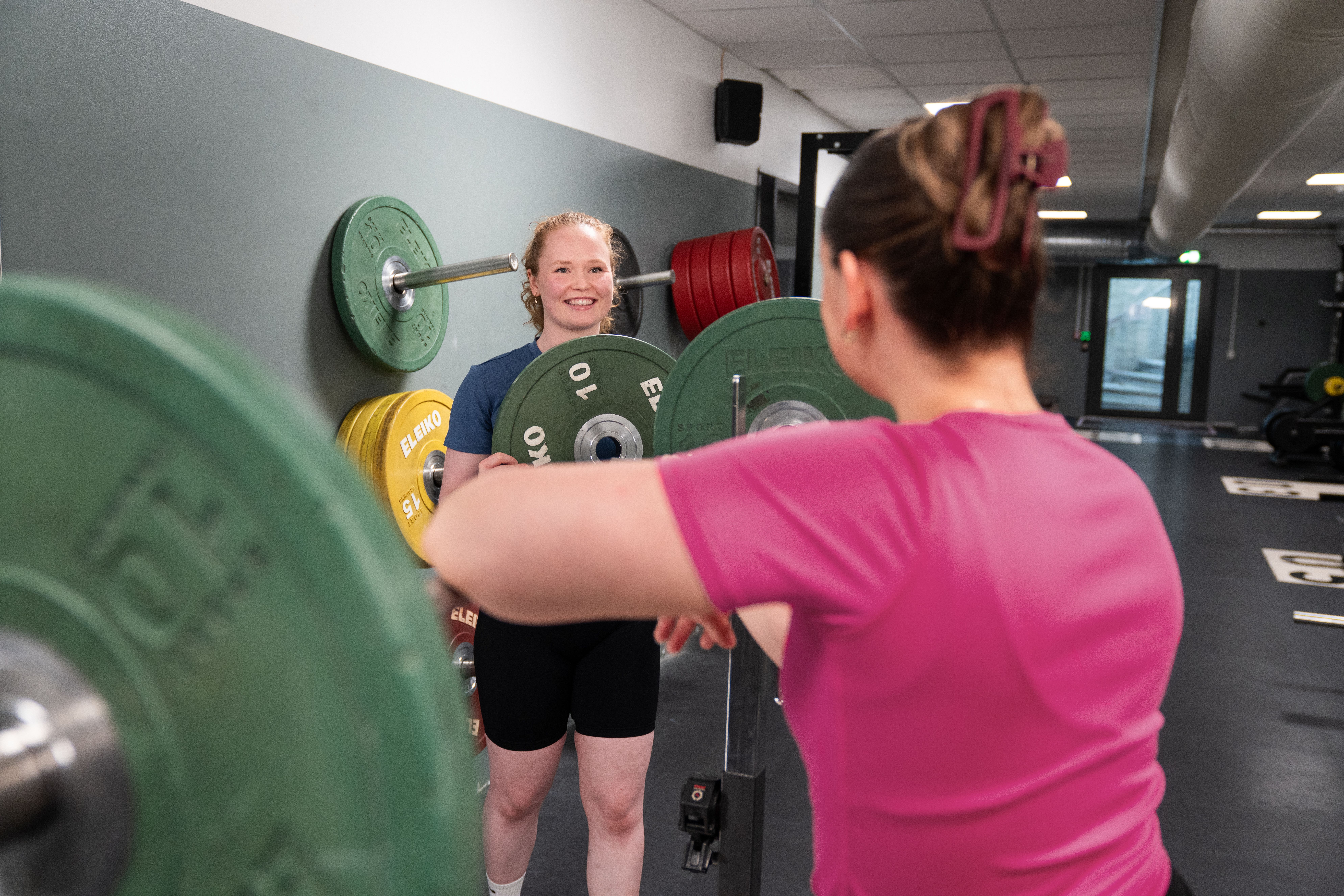 Personal trainer smiles and helps a student in a pink shirt to put weights on a barbell.