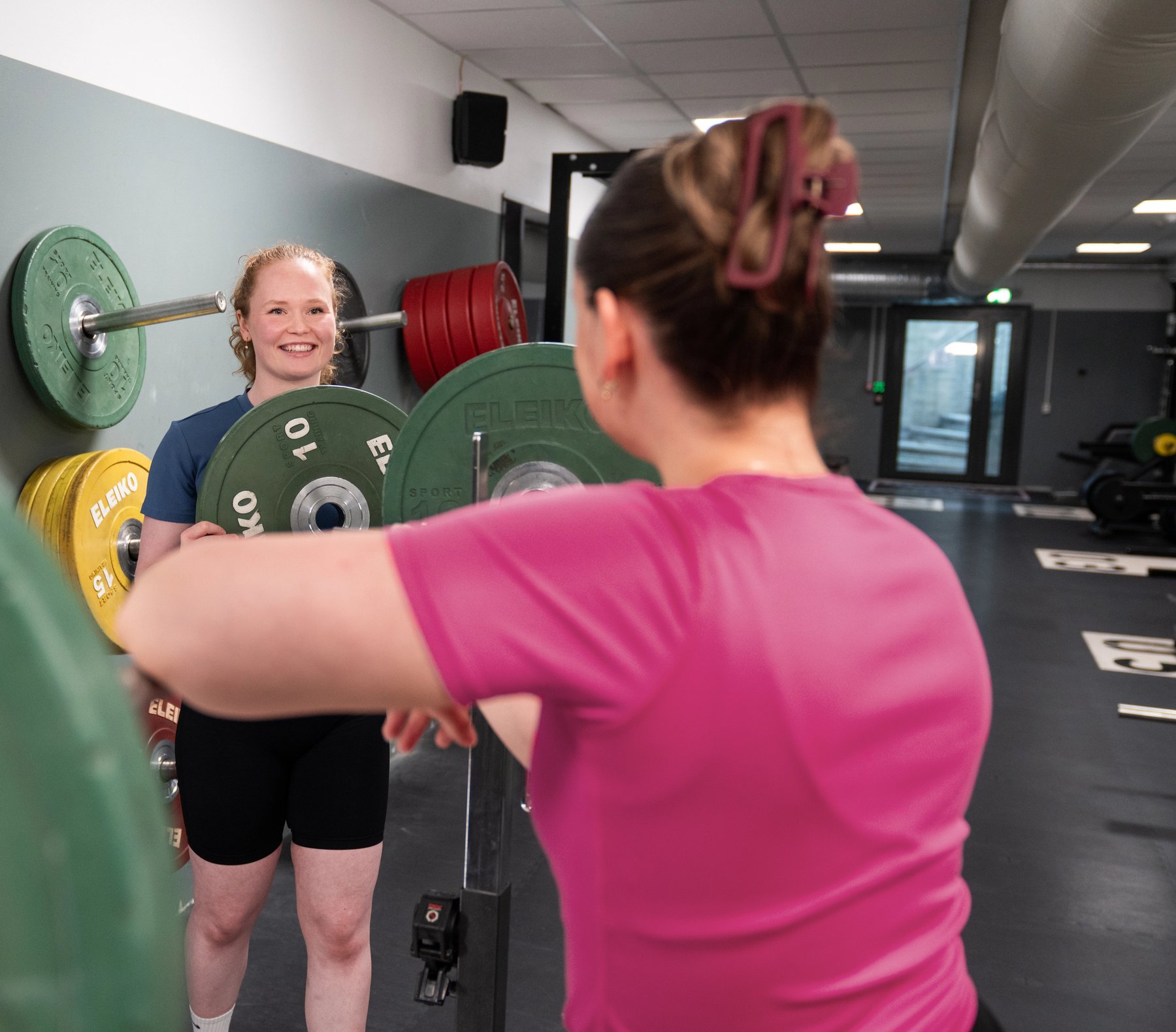 Personal trainer smiles and helps a student in a pink shirt to put weights on a barbell.