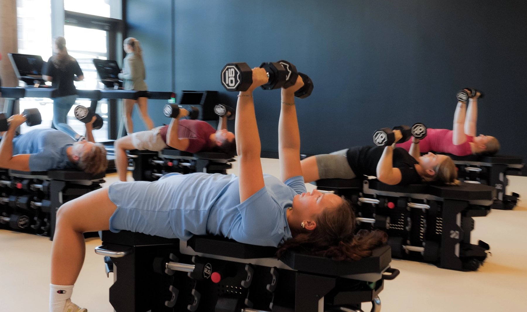 Flere unge mennesker som løper på tredemølle og trener styrke med manualer i en kombinasjonssal for løping og styrke.