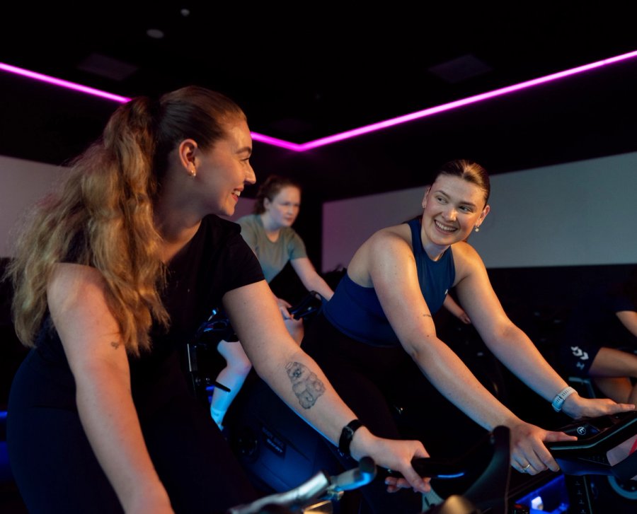 Two students are working out together on spinning bikes and smiling at each other in a Sit gym.