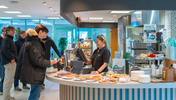 En student kjøper noe fra kantina på Gjøvik mens en gruppe studenter står bak ham og det står en smilende dame i kassa.