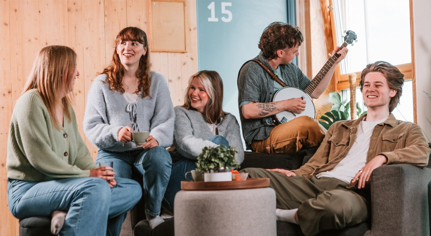 A group of students sitting together in a shared living room, with sofas, a small table and one person playing a string instrument.