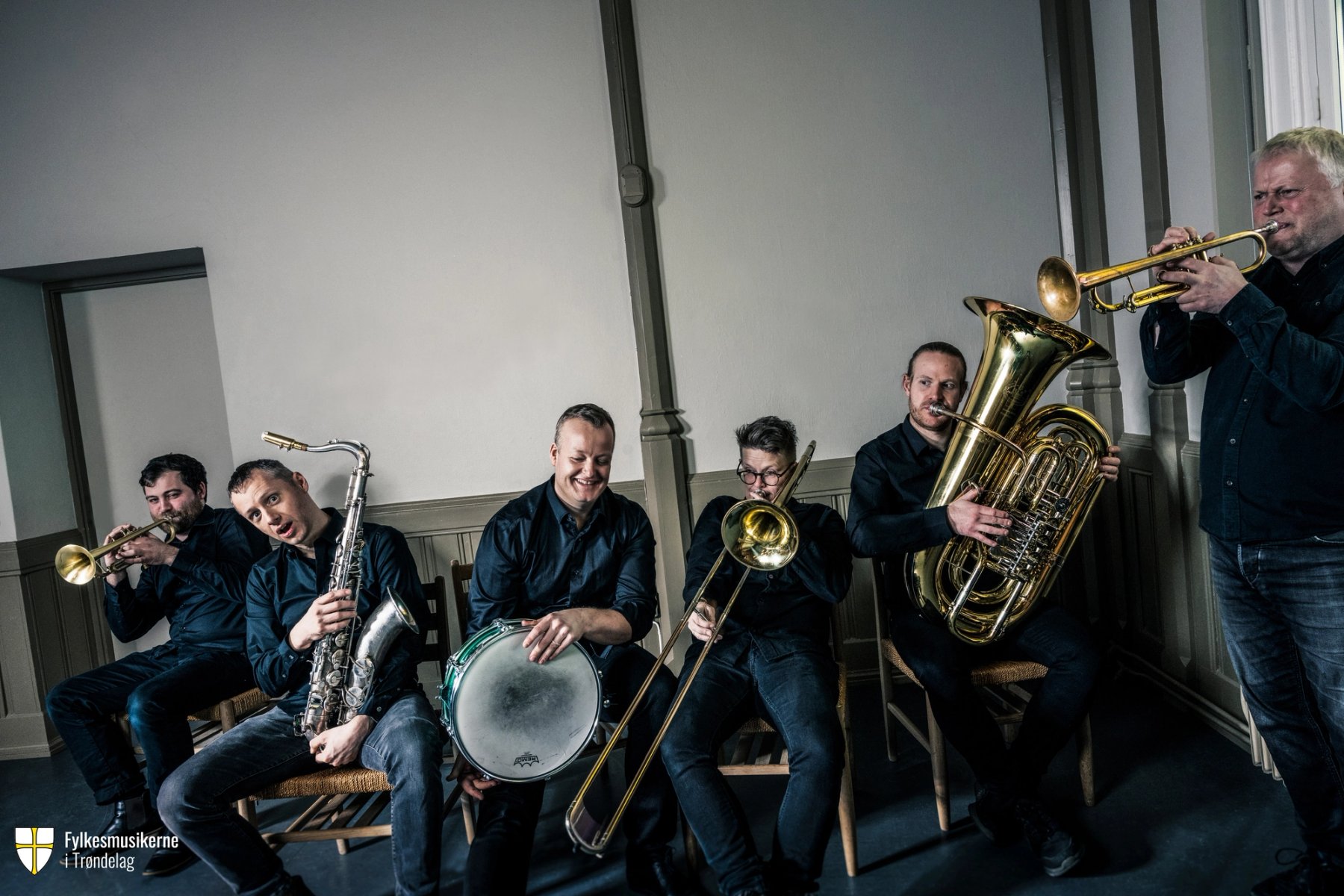 Seks musikere i bandet Quintus sitter på stoler og spiller ulike blåseinstrumenter og tromme, iført mørke skjorter, i et lyst rom.