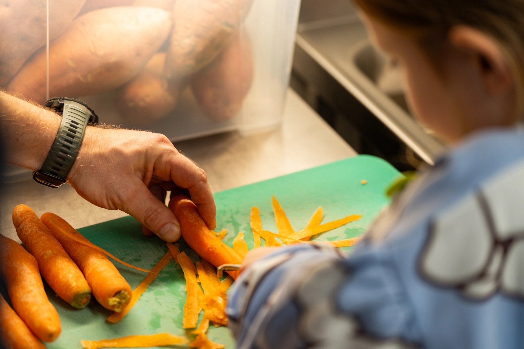 Photo of a child and an adult peeling carrots together in the kitchen. In the background, there is a plastic crate filled with sweet potatoes.