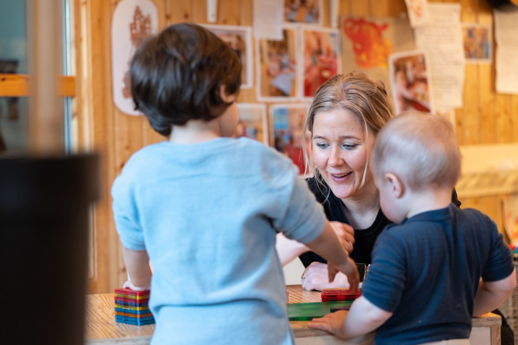 Photo of a kindergarten staff member smiling and engaging in play with two small children around a table, where they are building with colourful blocks. The walls are decorated with children's artwork and photos.