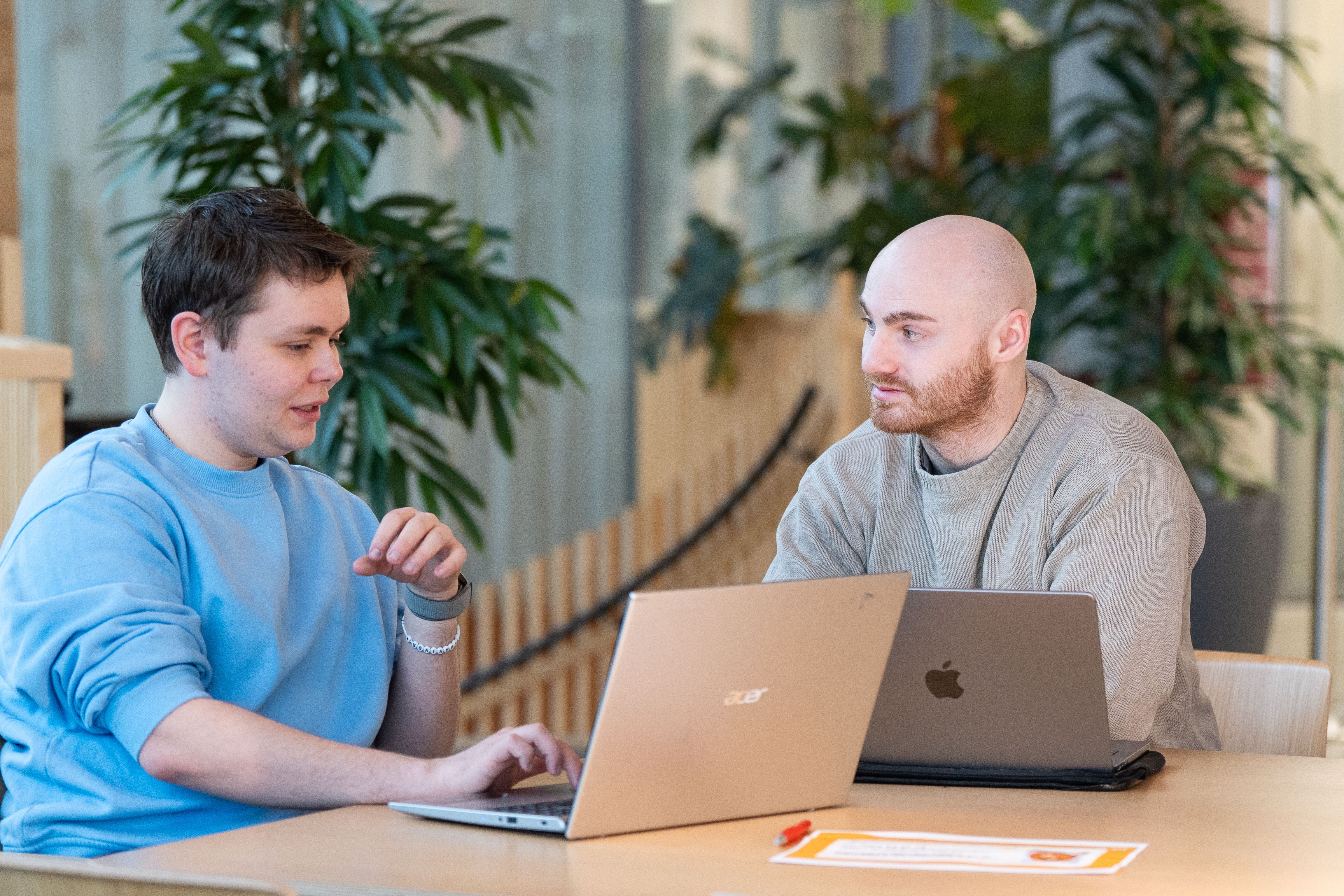 To studenter sitter ved et bord i et fellesareal og jobber sammen på hver sin bærbare datamaskin.