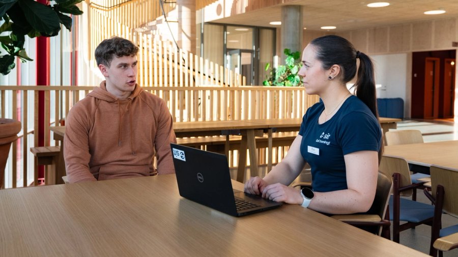 Photo of a physiotherapist talking with a young man at a table in a bright communal area, with a laptop in front of them.