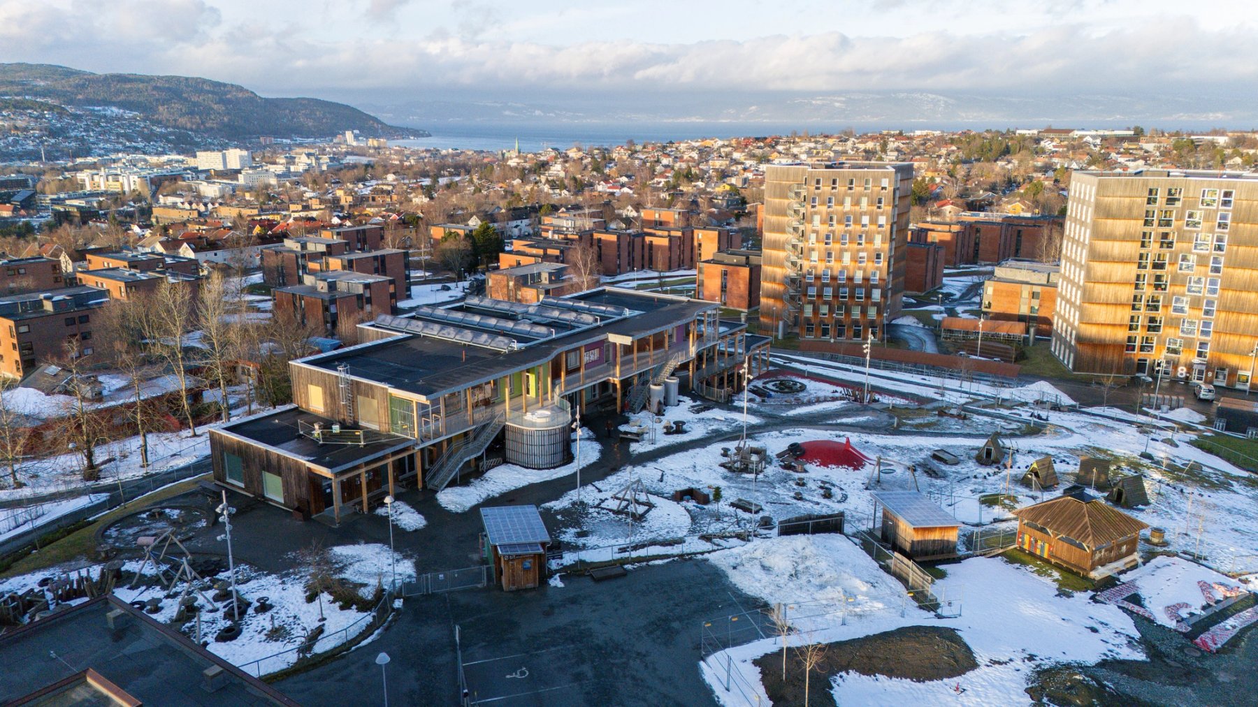 Drone photo of Moholt Student Village in winter. In the foreground is the kindergarten building with its play area, surrounded by student housing and snow-covered walkways. Trondheim and the fjord can be seen in the background.