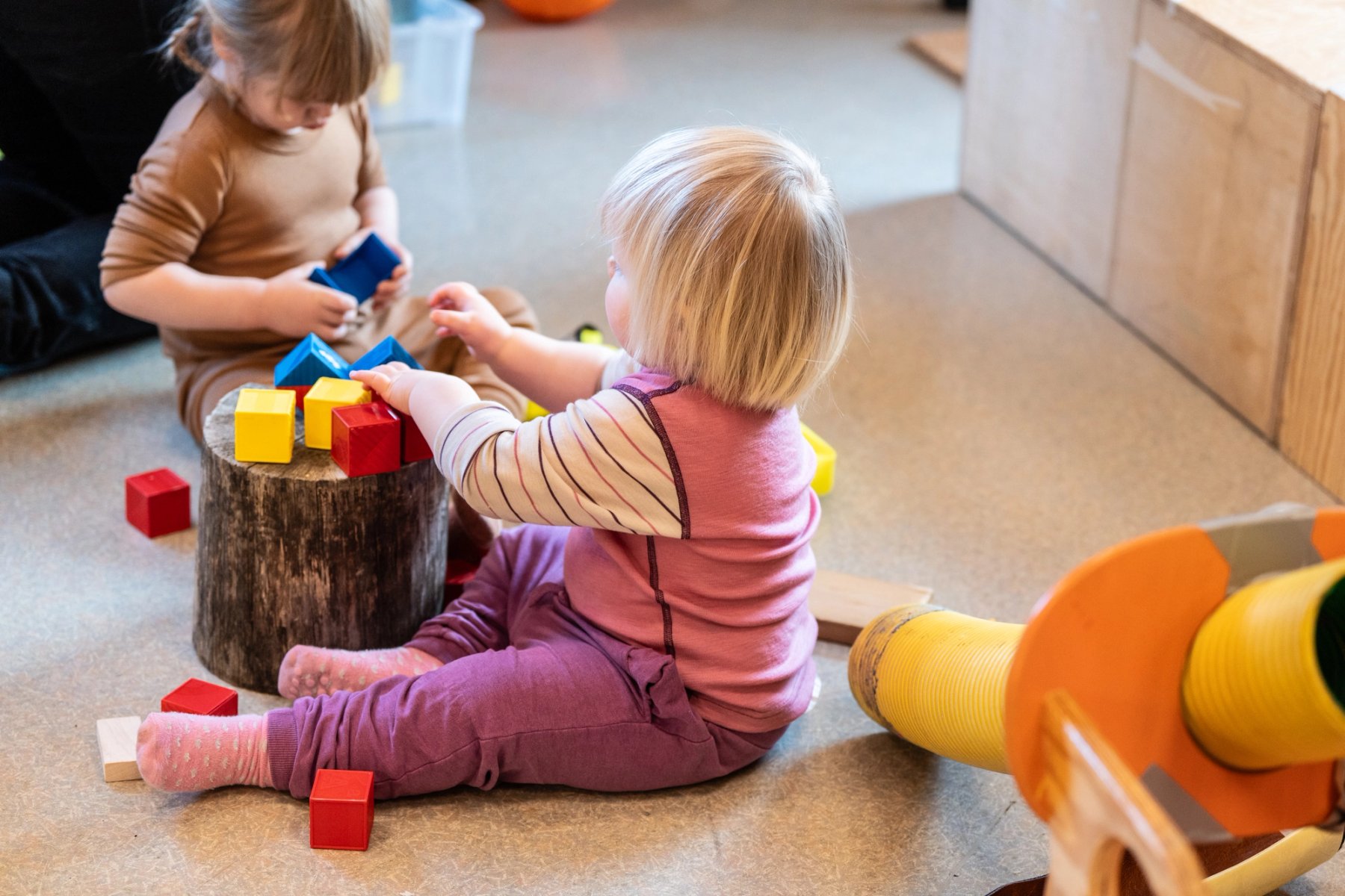 Picture of two small children who sit on the floor in the kindergarten, playing with colourful building blocks. A tree stump is used as a table, and the children are focused as they stack the blocks.