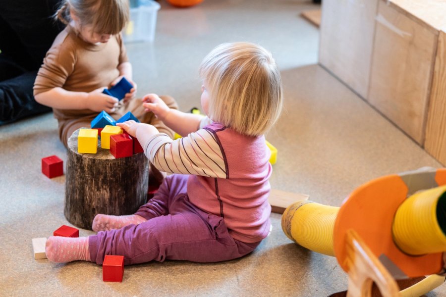 Photo of two small children sitting on the floor in the kindergarten, playing with colourful building blocks. A tree stump is used as a table, and the children are focused as they stack the blocks.