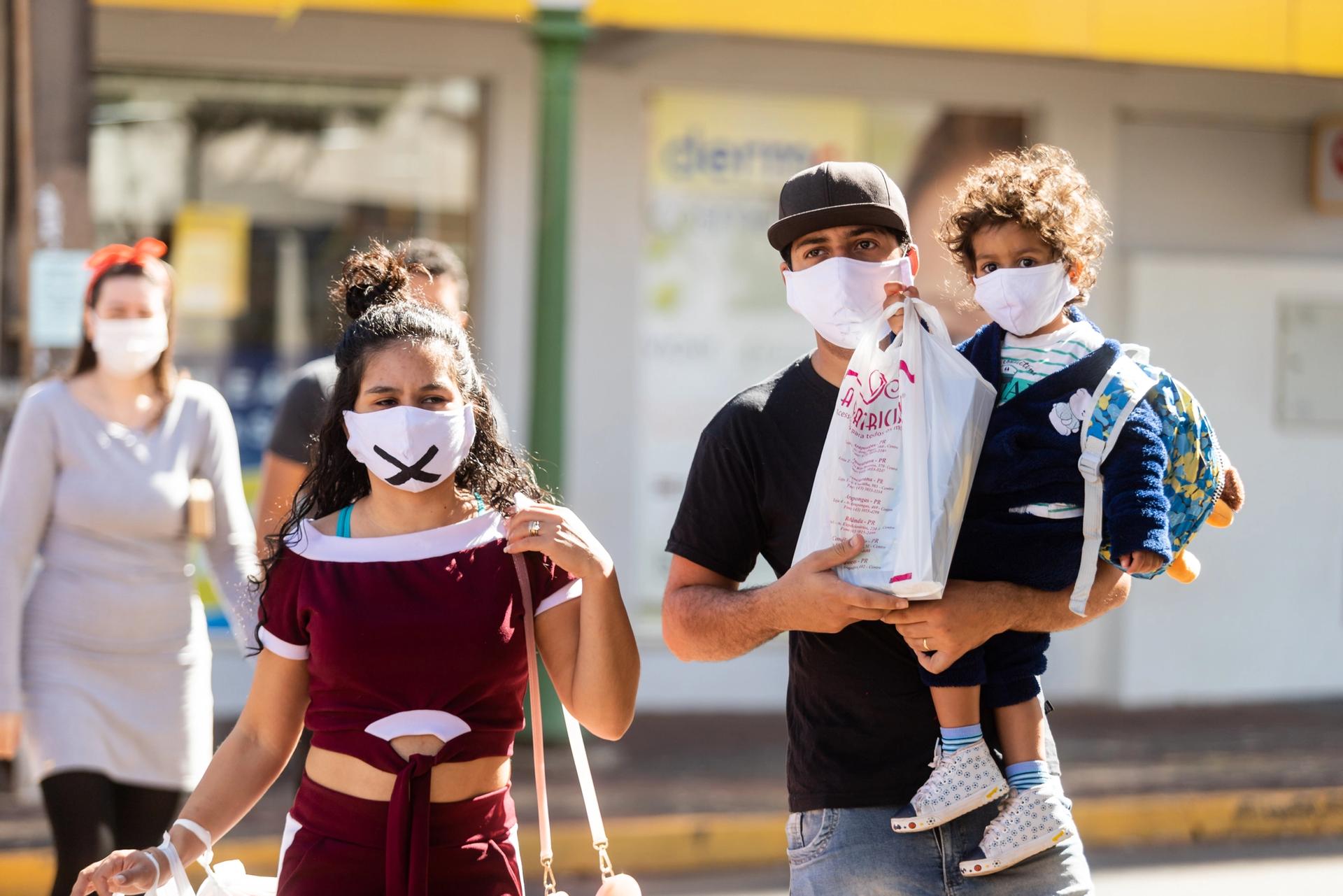 A woman, and a man carrying a young child, walking down the street towards the camera. All three are wearing masks over mouth and nose.