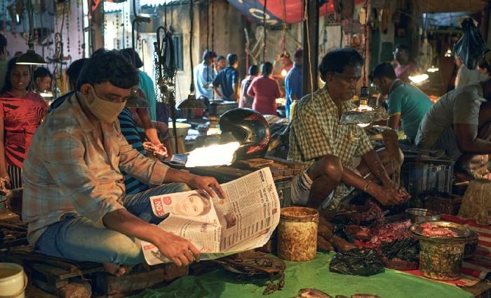 Inside a fish market, a fish trader is seen reading coronavirus related news in a Bengali newspaper, while other buyers and fish sellers are seen busy in their work.