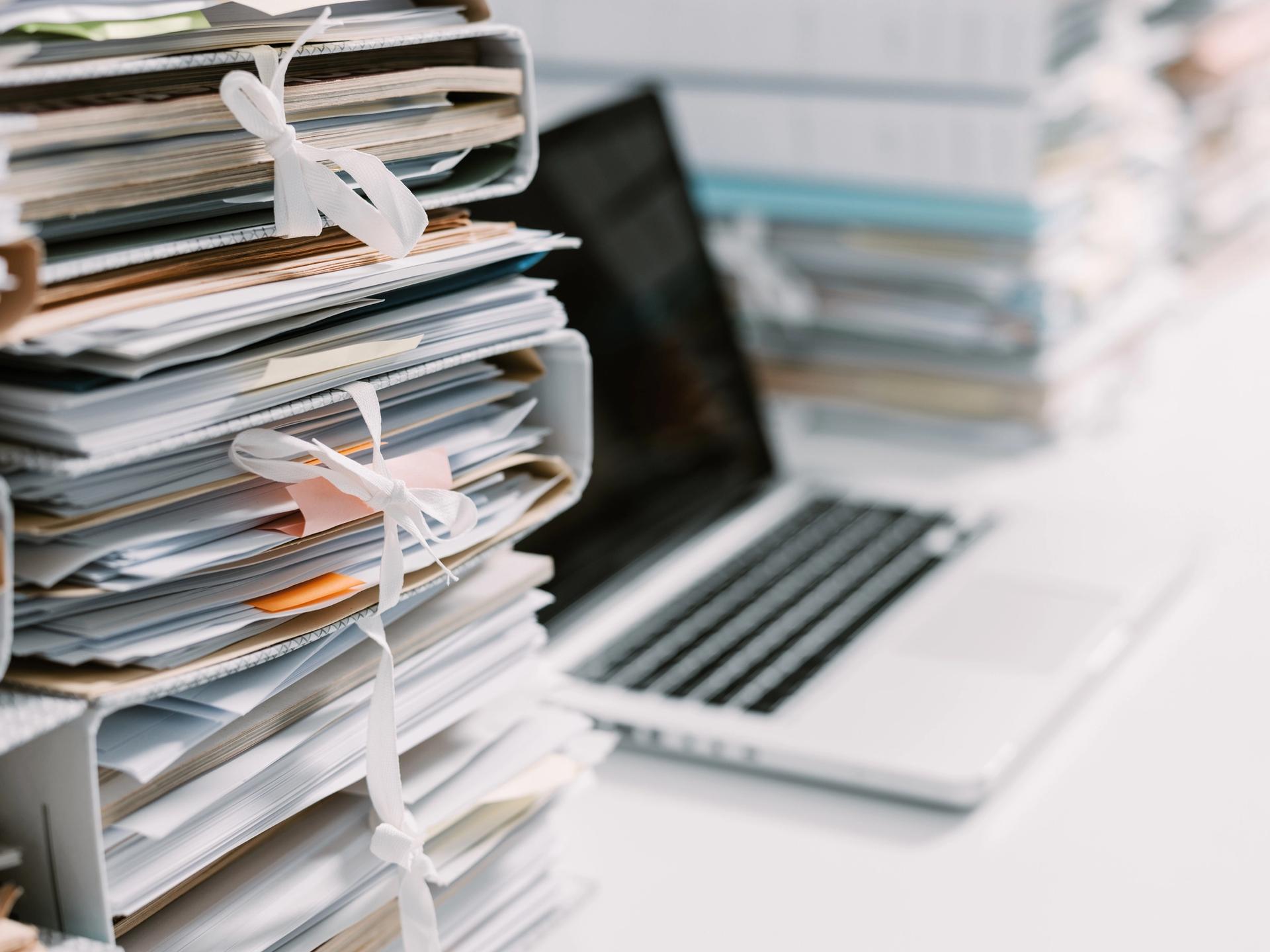 Photograph of a desk, with a pile of papers and folders in the foreground. A laptop with blank screen is behind them.