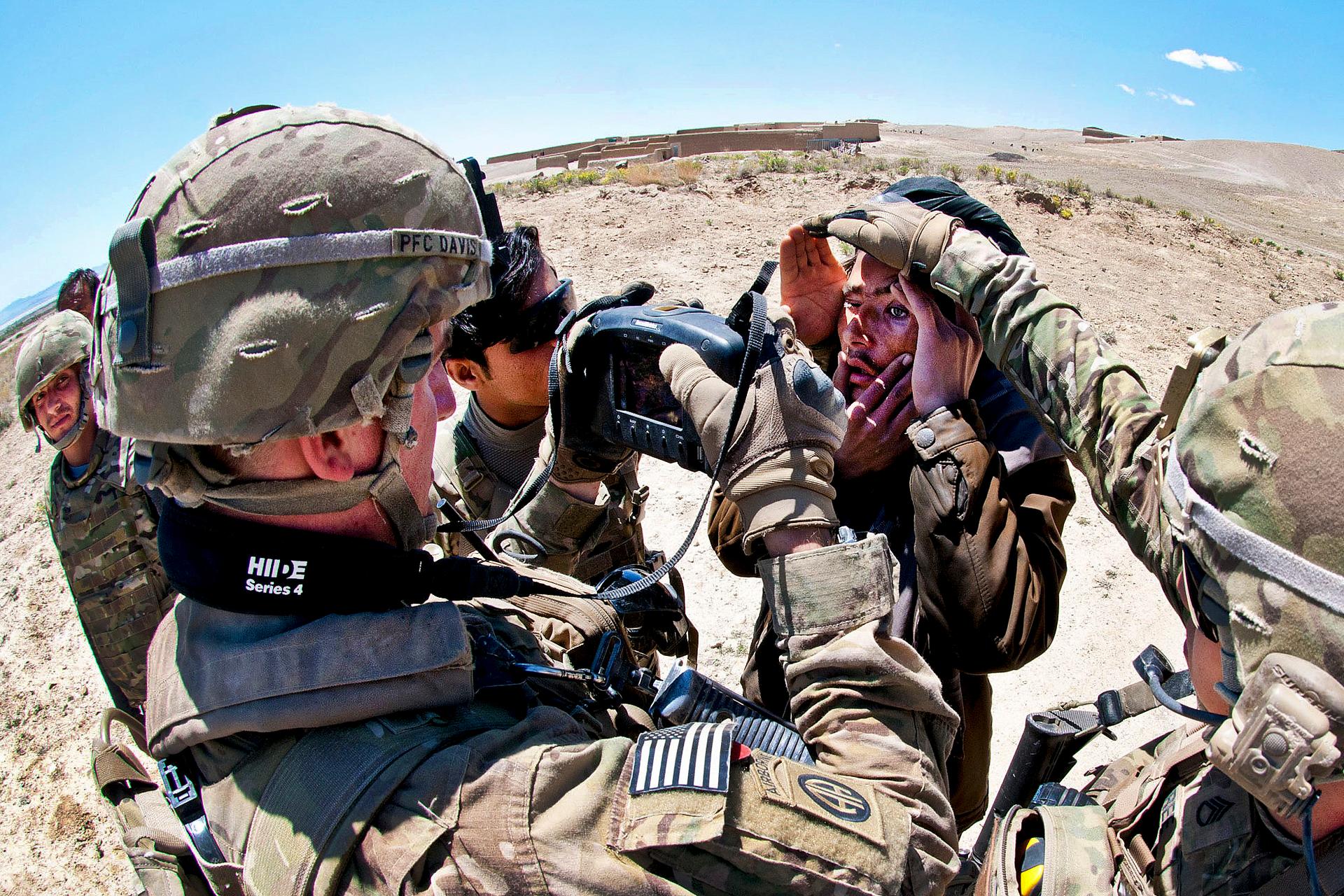 U.S. paratroopers scan the eye of an Afghan civilian. One soldier shields the young man's eyes from the sun while another uses a scanner..