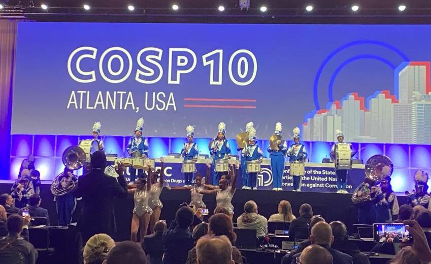 Photo of a marching band on stage at a conference. The large screen behind the performers reads "COSP10 Atlanta, USA"