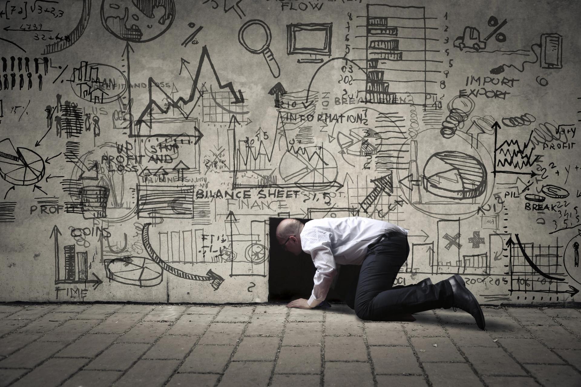 A man on hands and knees, crawling into a small gap in a wall covered with business-related sketches.
