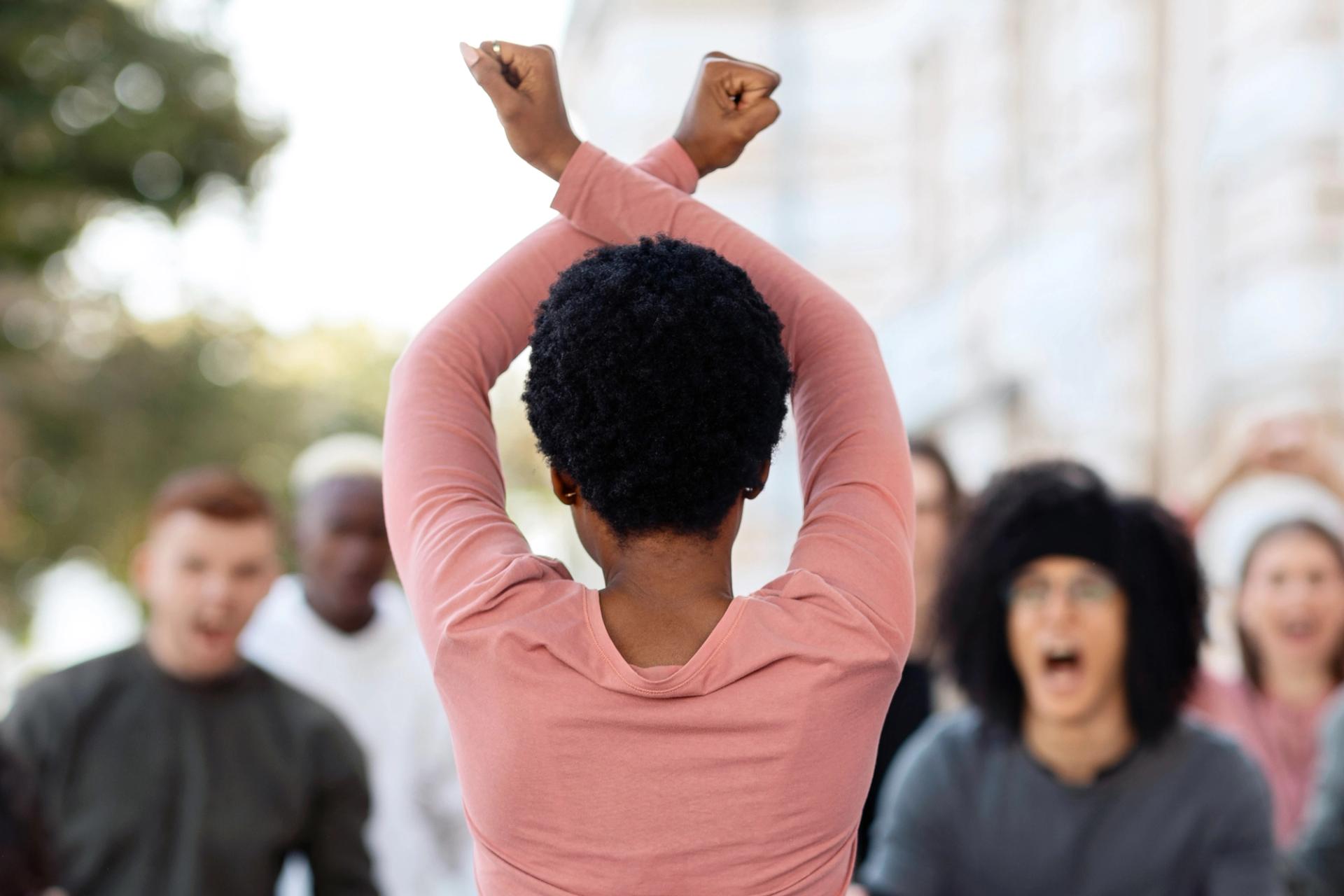 Rear-view photograph of a woman of African heritage with arms raised above her head in a cross shape. Blurred faces can be seen in the background, facing the woman and looking angry.