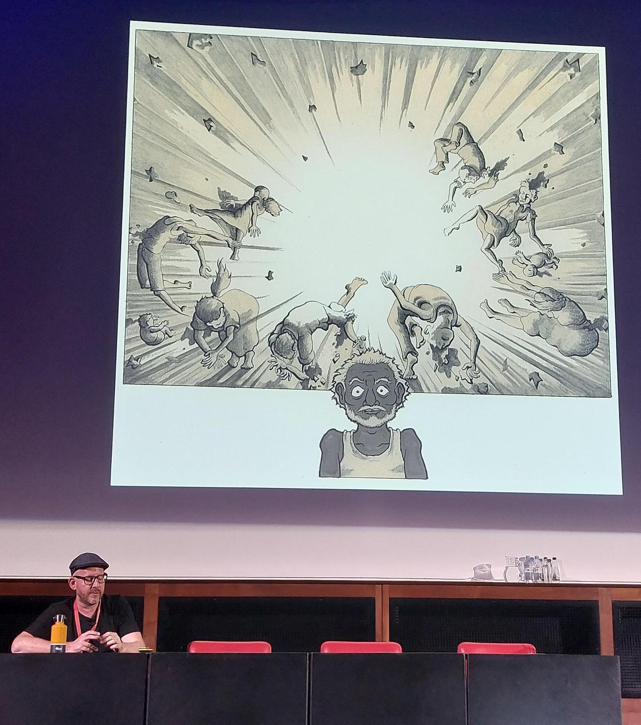 A panelist sits beneath a large screen showing a single panel from the graphic novel. The illustration shows a man in front of an explosion.