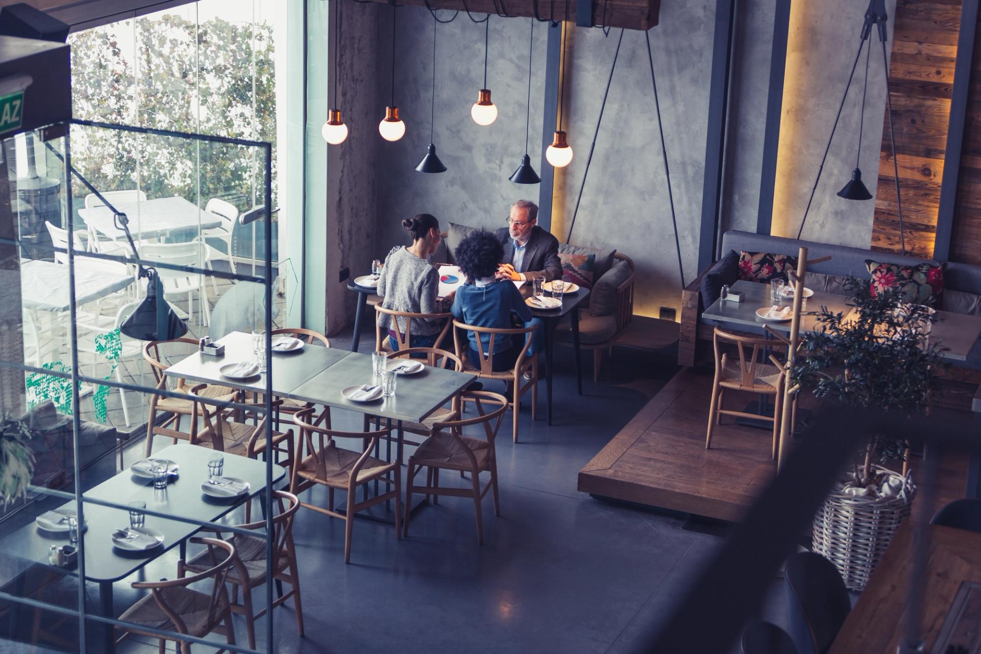 A photograph of the interior of a modern restaurant. Three people are sat at a table, having a business meeting. Two people have their backs to the camera; one man is facing them across the table.