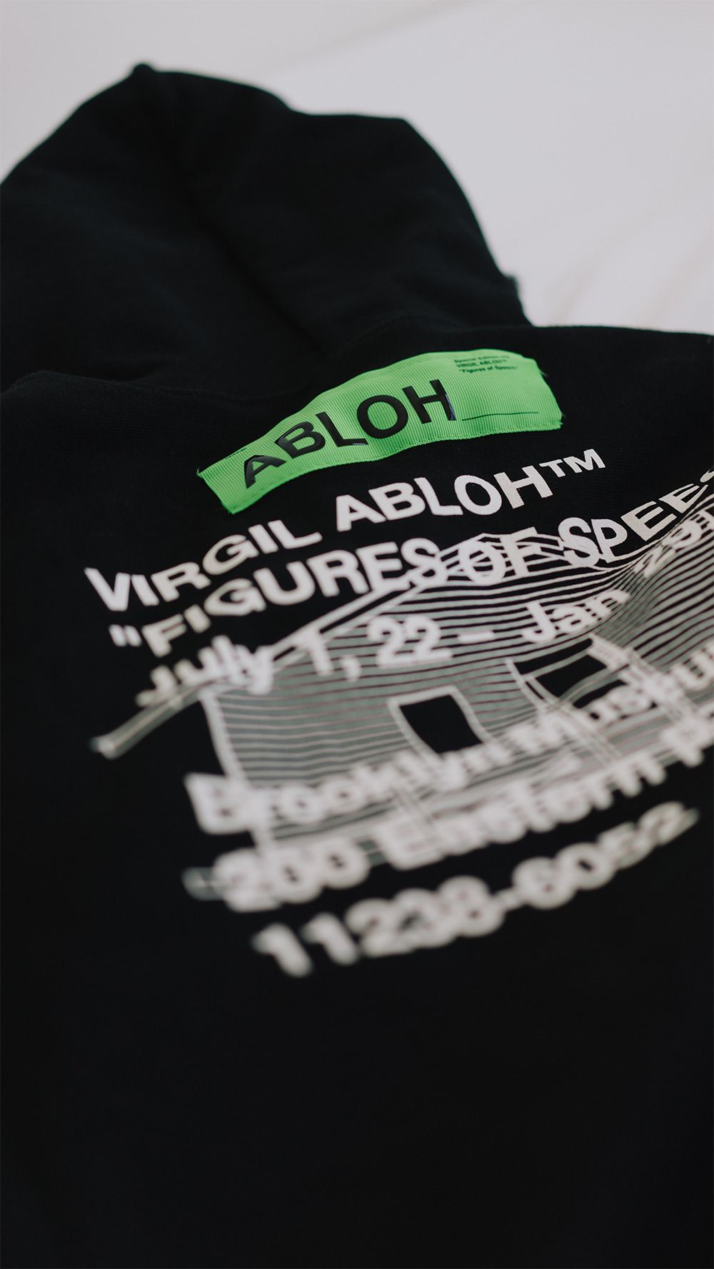 Black garment with neon green "ABLOH" and white text reading "VIRGIL ABLOH™ FIGURES OF SPEE" against plain white background, featuring blurred dates "July 7, 22 – Jan" and building-like graphic with windows, faint additional text and numbers at base.
