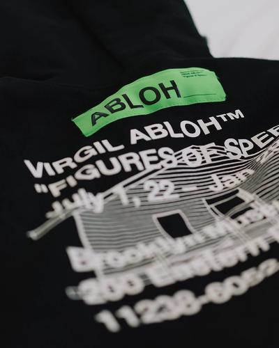 Black garment with neon green "ABLOH" and white text reading "VIRGIL ABLOH™ FIGURES OF SPEE" against plain white background, featuring blurred dates "July 7, 22 – Jan" and building-like graphic with windows, faint additional text and numbers at base.